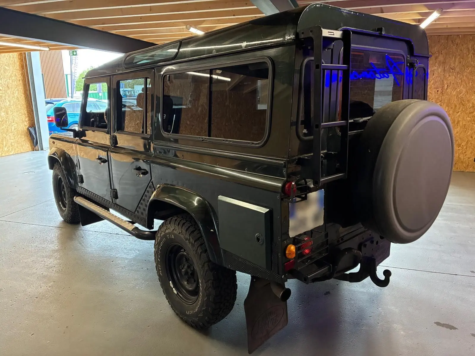 Land Rover Defender 110 vert vue 3/4 arrière droit, avec roue de secours et échelle sur porte arrière.