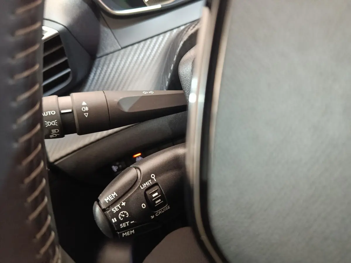 Gros plan sur les commandes du régulateur de vitesse et des feux, côté gauche du volant cuir noir de la Peugeot 208 blanche.
