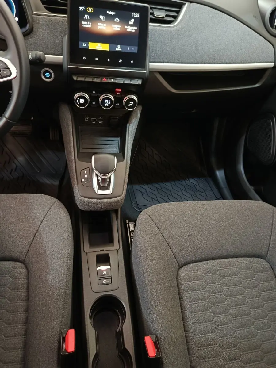 Vue intérieure centrée sur la console centrale et les sièges gris de la Renault ZOE électrique blanche, avec écran tactile et levier de vitesses.