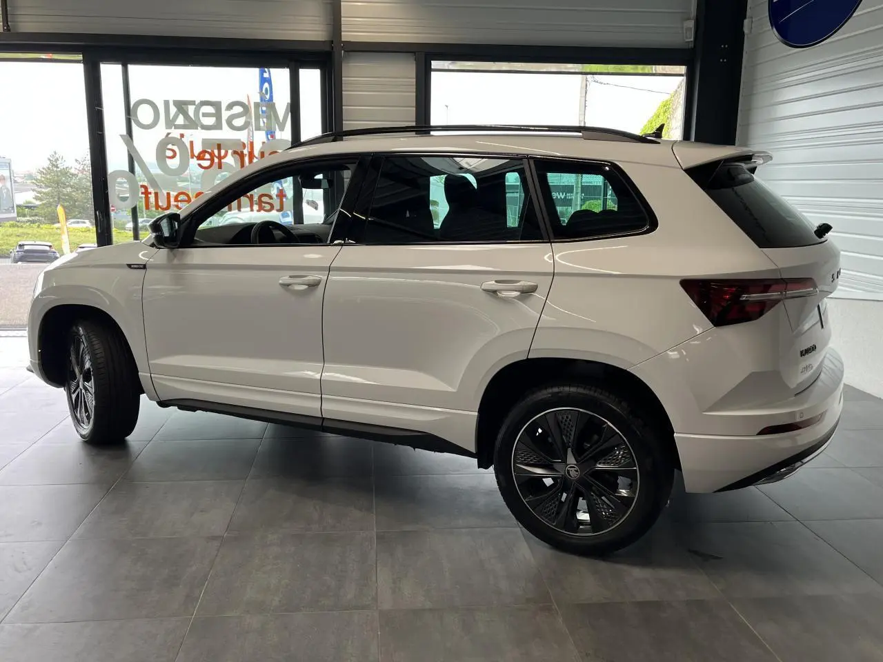 Profil droit du SUV SKODA KAROQ blanc 2025 avec jantes alliage noires et toit ouvrant visible en intérieur showroom.