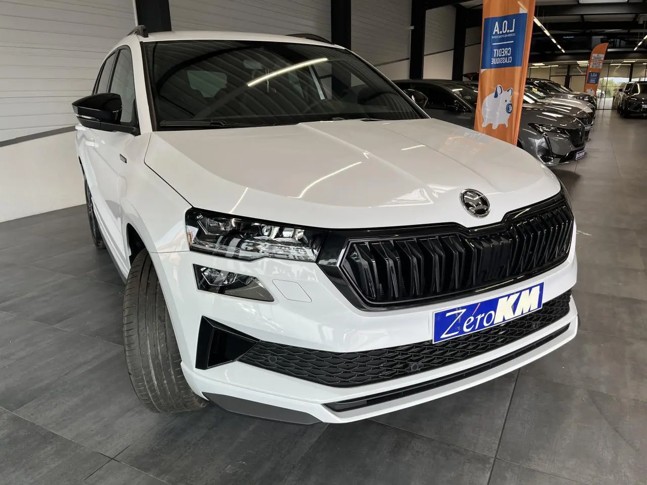 Vue 3/4 avant d'un SUV Skoda Karoq blanc avec calandre noire et phares LED, roues braquées à gauche en showroom.