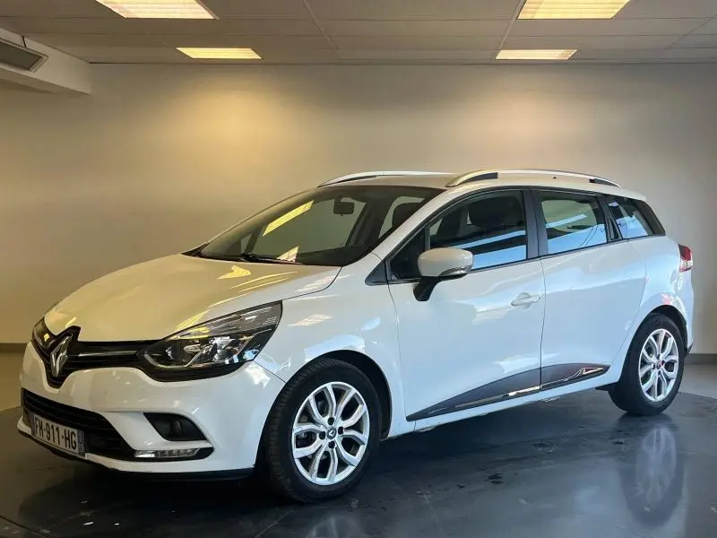 Renault Clio Estate blanc glacier vue 3/4 avant droit en intérieur avec barres de toit et jantes alliage.