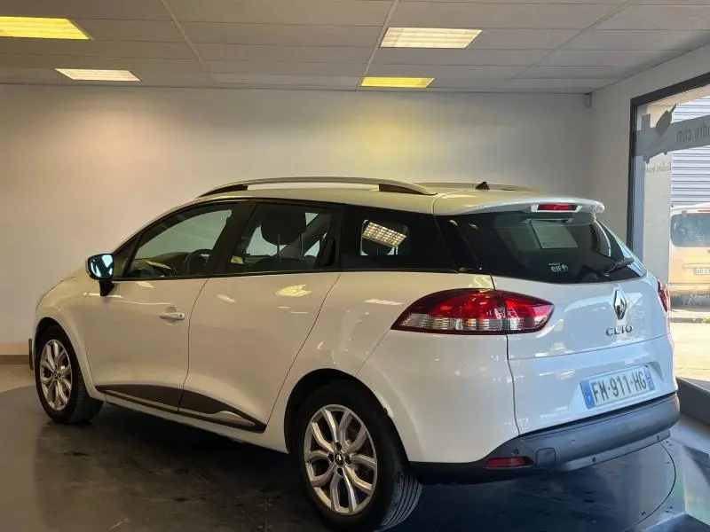Renault Clio Estate blanc Glacier vue 3/4 arrière droit avec barres de toit et becquet arrière dans un showroom.