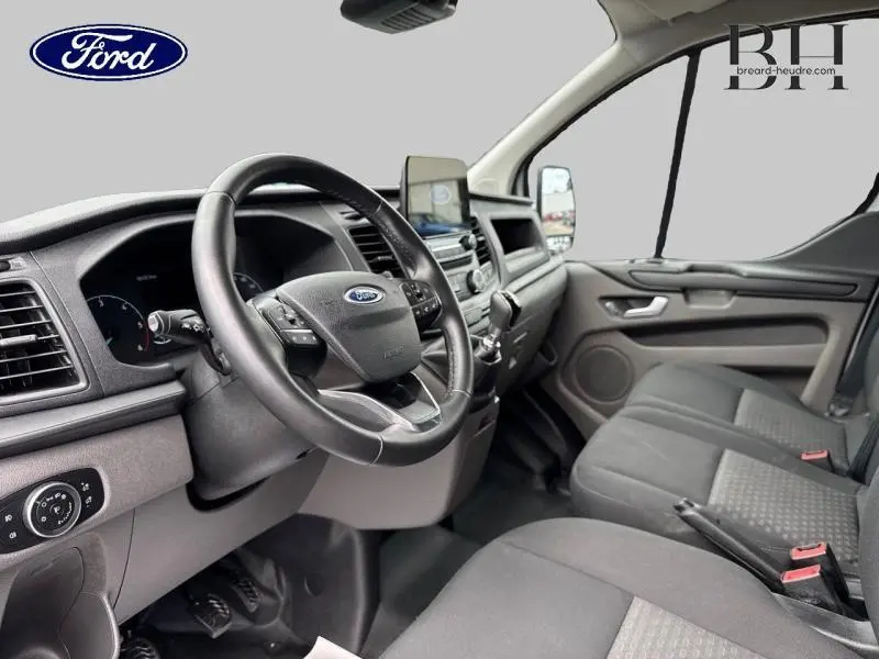 Intérieur du Ford Transit Custom blanc, vue côté conducteur montrant le volant cuir et la banquette avant grise.