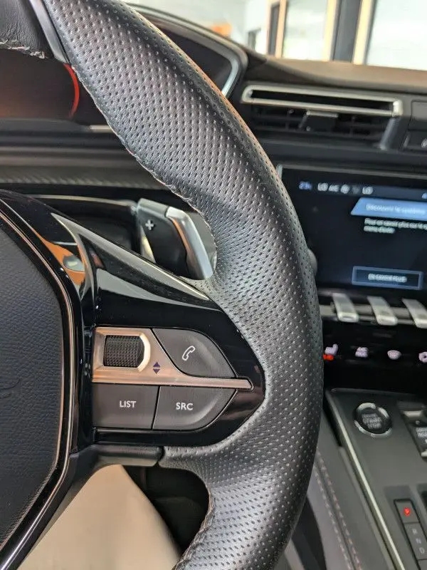 Gros plan sur le volant cuir perforé noir de la Peugeot 508 SW 2024 avec commandes audio et tableau de bord gris noir.