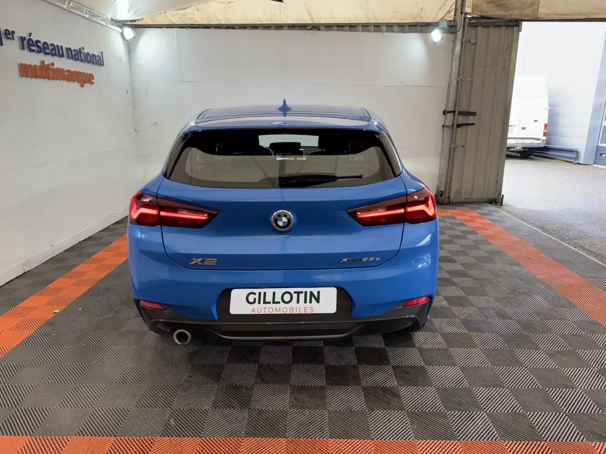 Vue arrière d’un BMW X2 xDrive25e M Sport bleu, avec feux allumés et sortie d’échappement visible côté gauche.