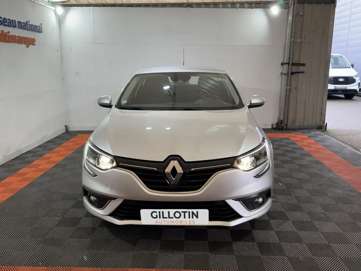 Vue avant d'une Renault Mégane Business gris clair avec phares allumés dans un garage éclairé.