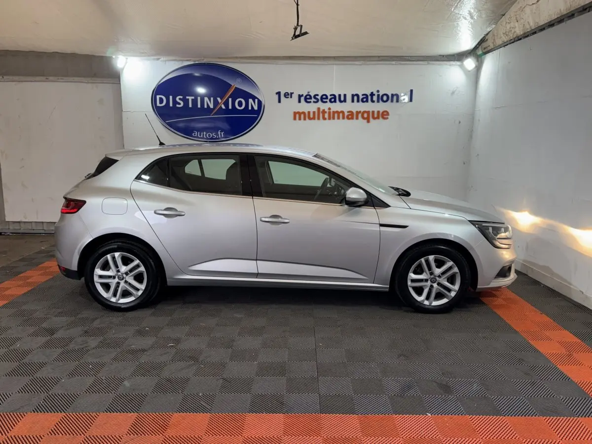 Profil droit d'une Renault Mégane Business gris clair, stationnée en intérieur sur sol noir et orange.