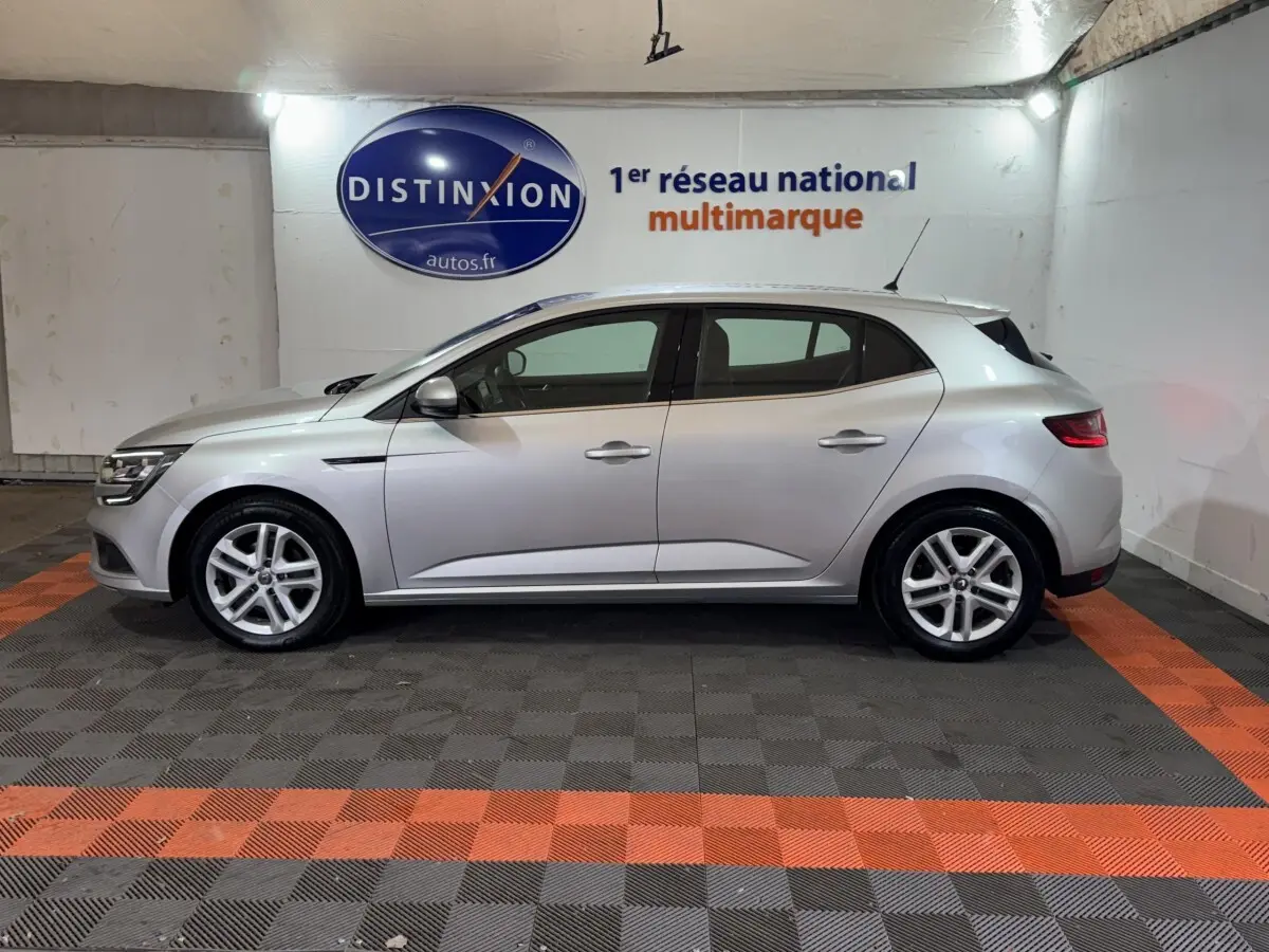 Profil droit d'une Renault Mégane Business gris clair, en intérieur sur sol à damiers gris et orange, avec logo Distinxion en arrière-plan.