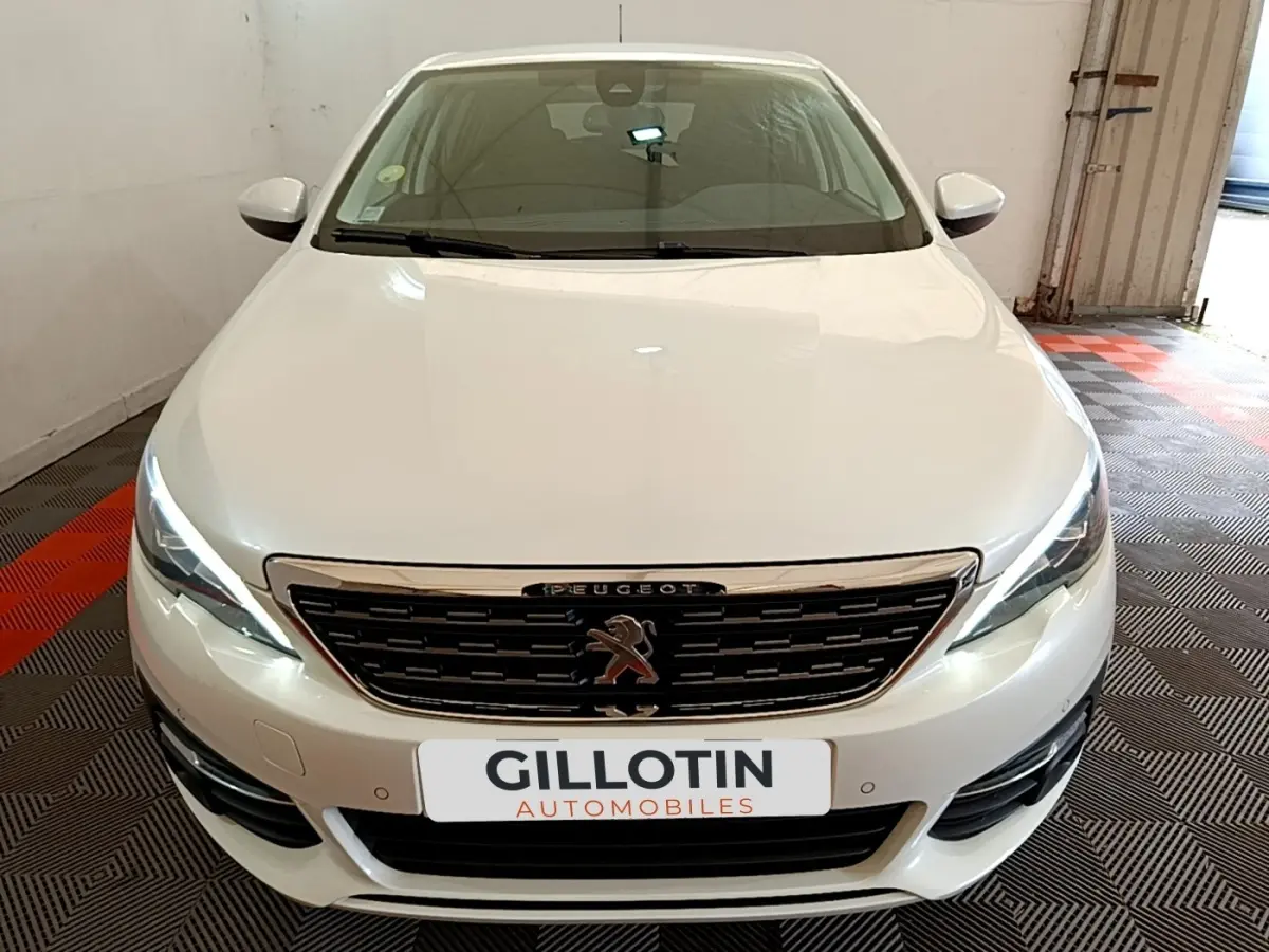 Vue avant frontale d'une Peugeot 308 blanche avec calandre chromée et feux LED allumés dans un garage.