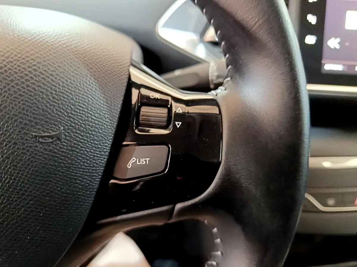 Gros plan sur les commandes du volant cuir noir de la Peugeot 308, avec écran tactile en arrière-plan.