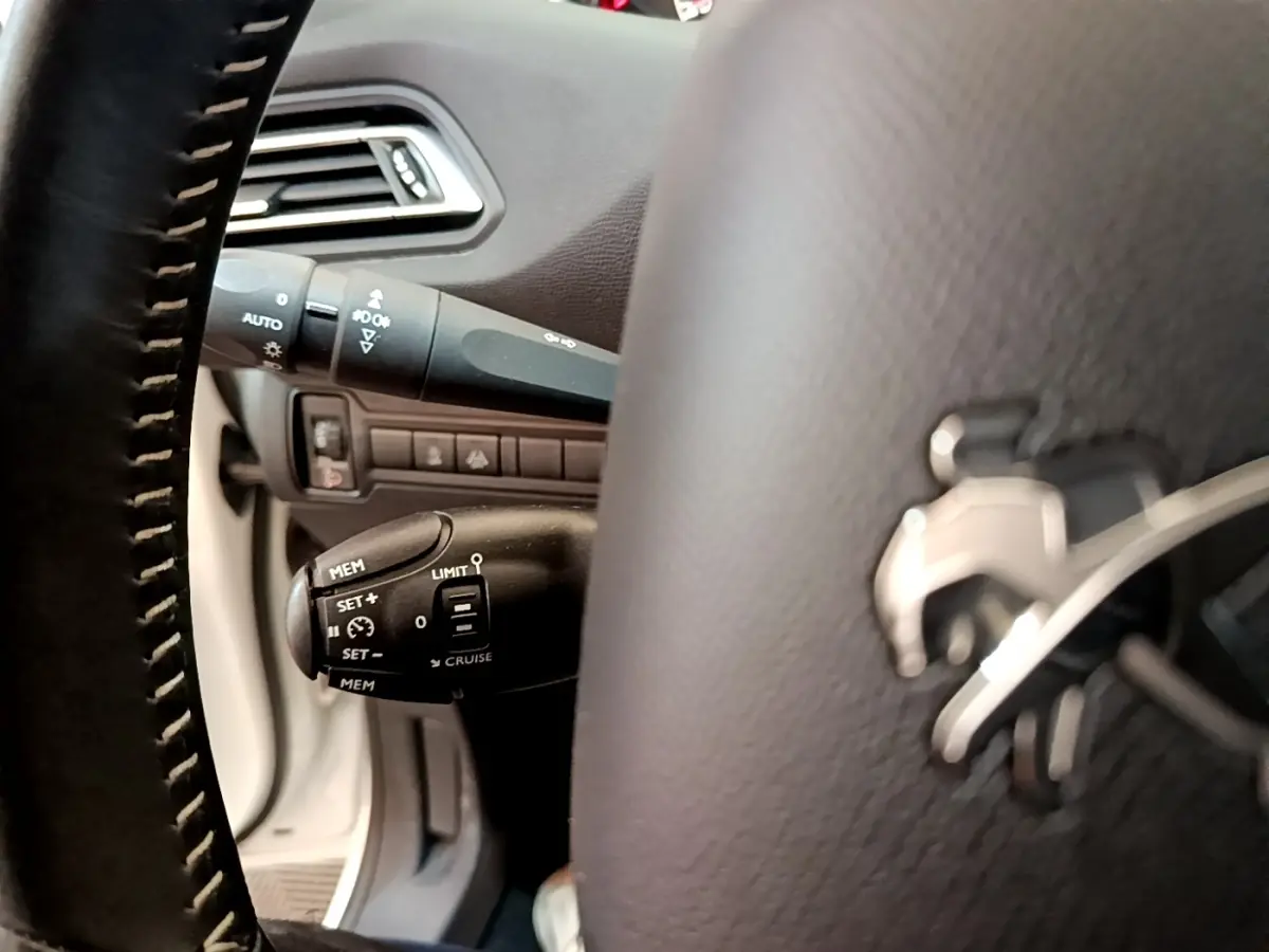 Gros plan sur le volant cuir noir de la Peugeot 308 blanc, avec commandes de régulateur de vitesse visibles à gauche.