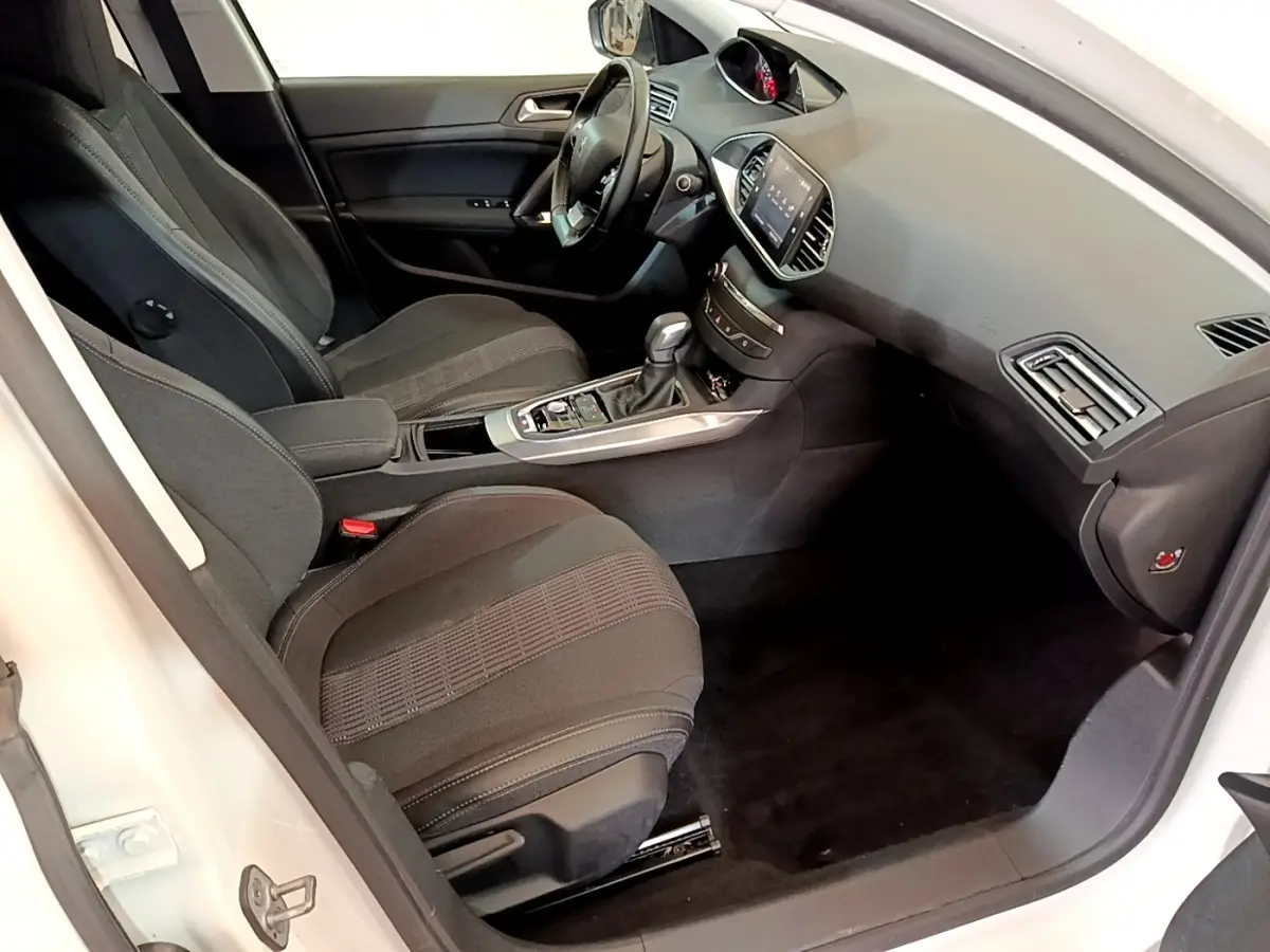 Vue intérieure côté conducteur de la Peugeot 308 blanche, montrant sièges noirs et tableau de bord avec écran tactile.