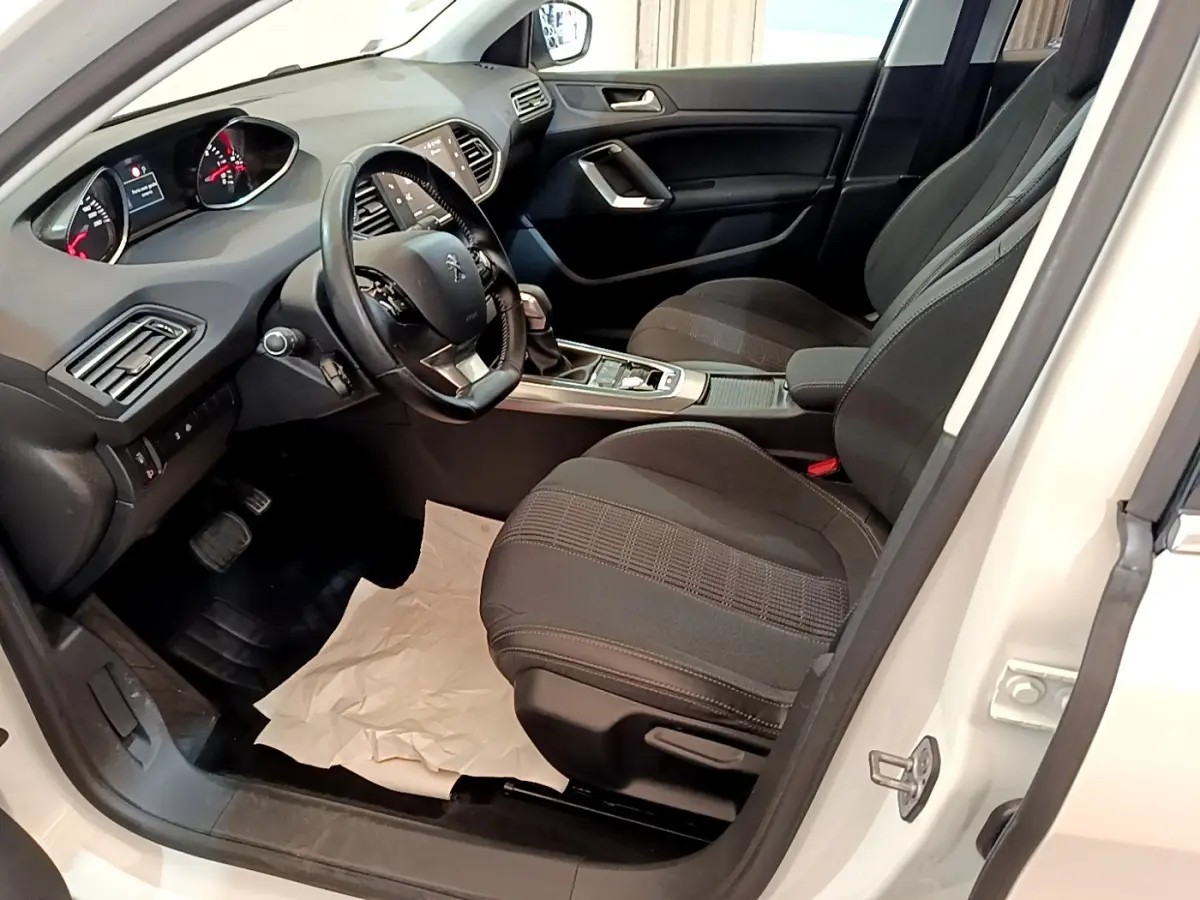 Vue intérieure côté conducteur de la Peugeot 308 blanche, montrant sièges noirs et tableau de bord avec écran tactile.