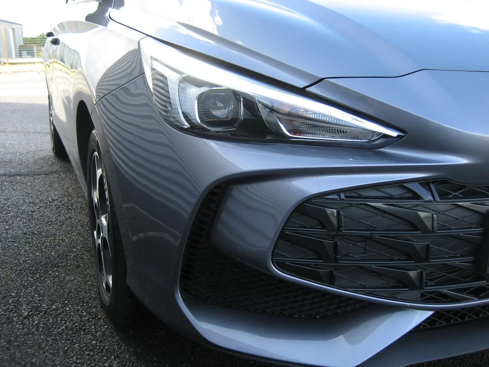Gros plan sur l'avant droit gris du MG3 1.5 Hybrid 195CV Luxury, mettant en valeur le phare et la calandre ajourée.