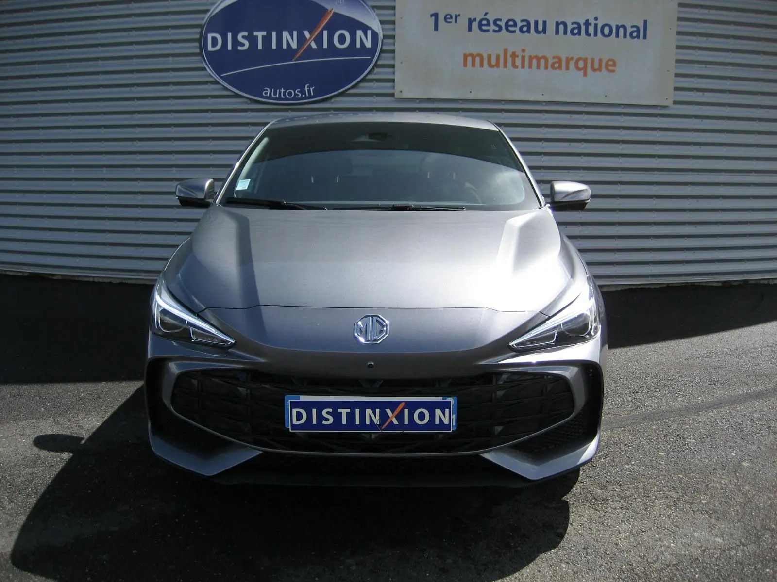 Vue de face d'une MG3 gris métallisé 2025 avec calandre noire et phares LED, devant un mur avec enseigne Distinxion.