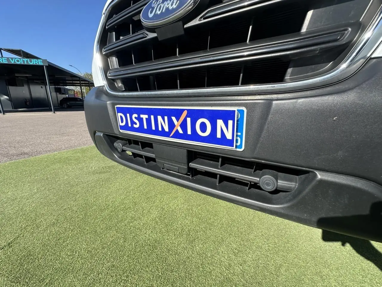 Gros plan sur la calandre avant noire et la plaque "DISTINXION" du Ford Transit Fourgon blanc de 2020.