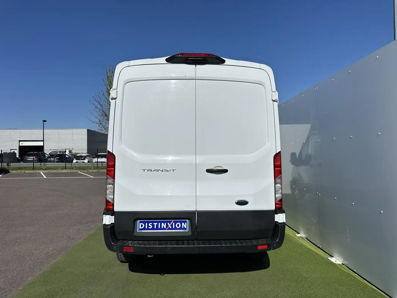 Vue arrière d'un fourgon Ford Transit blanc avec porte double et pare-chocs noir sous un ciel bleu.