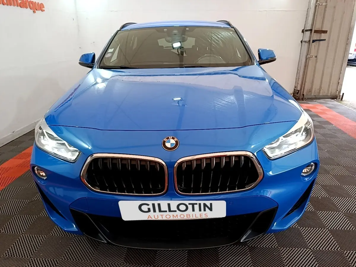 Vue avant d'un BMW X2 sDrive20i M Sport bleu, avec calandre noire et phares LED allumés, en intérieur showroom.