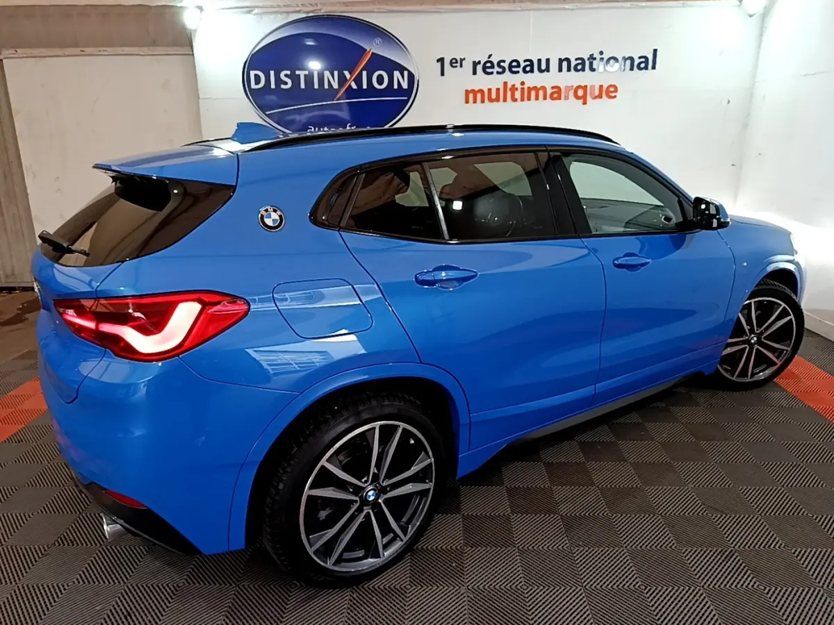 BMW X2 sDrive20i M Sport bleu en vue 3/4 arrière droit, avec jantes alliage et feux arrière allumés.