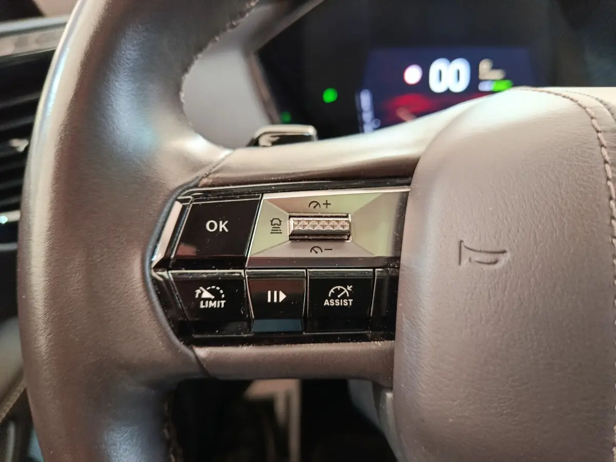 Gros plan sur les commandes gauche du volant cuir gris du DS4 E-TENSE 225 LA PREMIERE, affichage numérique en arrière-plan flou.