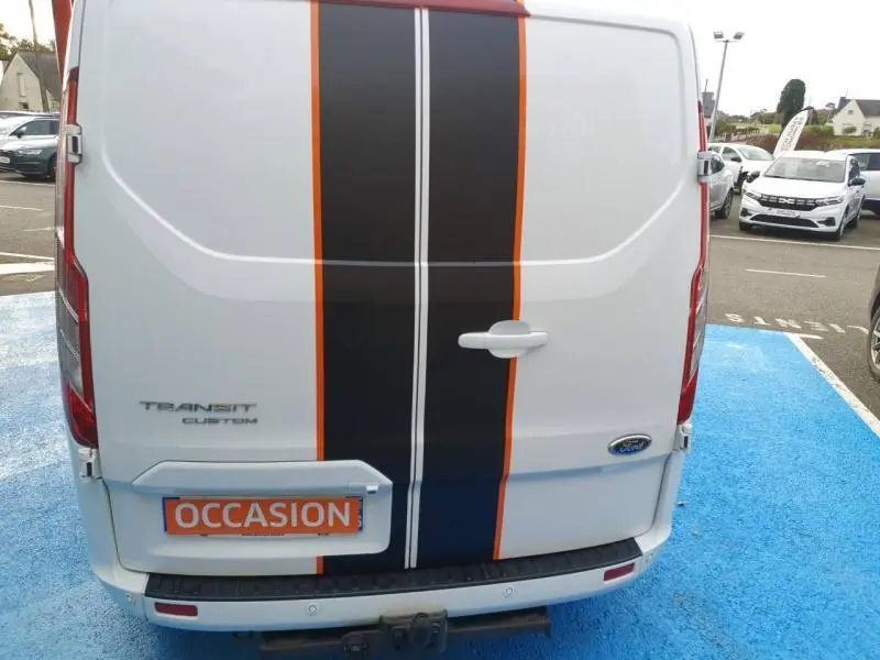 Vue arrière d'un Ford Transit Custom blanc glacier avec bandes noires et orange et crochet d'attelage visible.