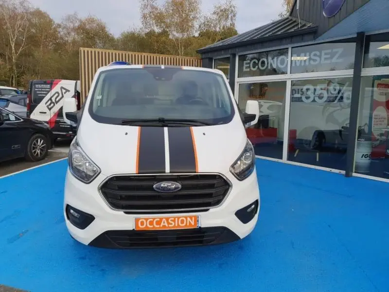 Vue avant d'un Ford Transit Custom blanc glacier avec bandes noires et orange sur le capot, sur parking bleu devant un magasin.