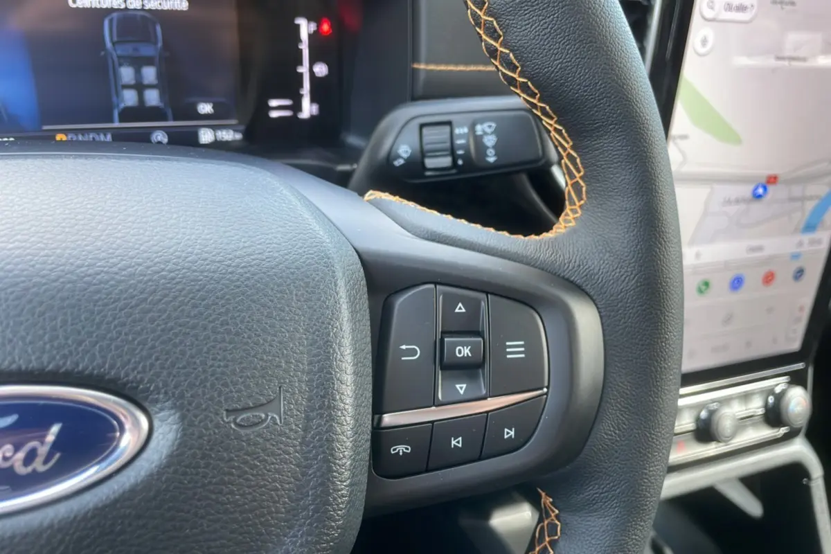 Gros plan sur le volant cuir noir avec surpiqûres orange du Ford Ranger Wildtrak 2025, affichage numérique et écran tactile visibles.