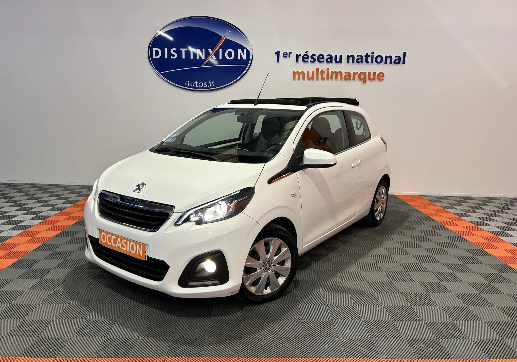 Peugeot 108 blanc vue 3/4 avant droit avec toit ouvrant et phares allumés en intérieur showroom.