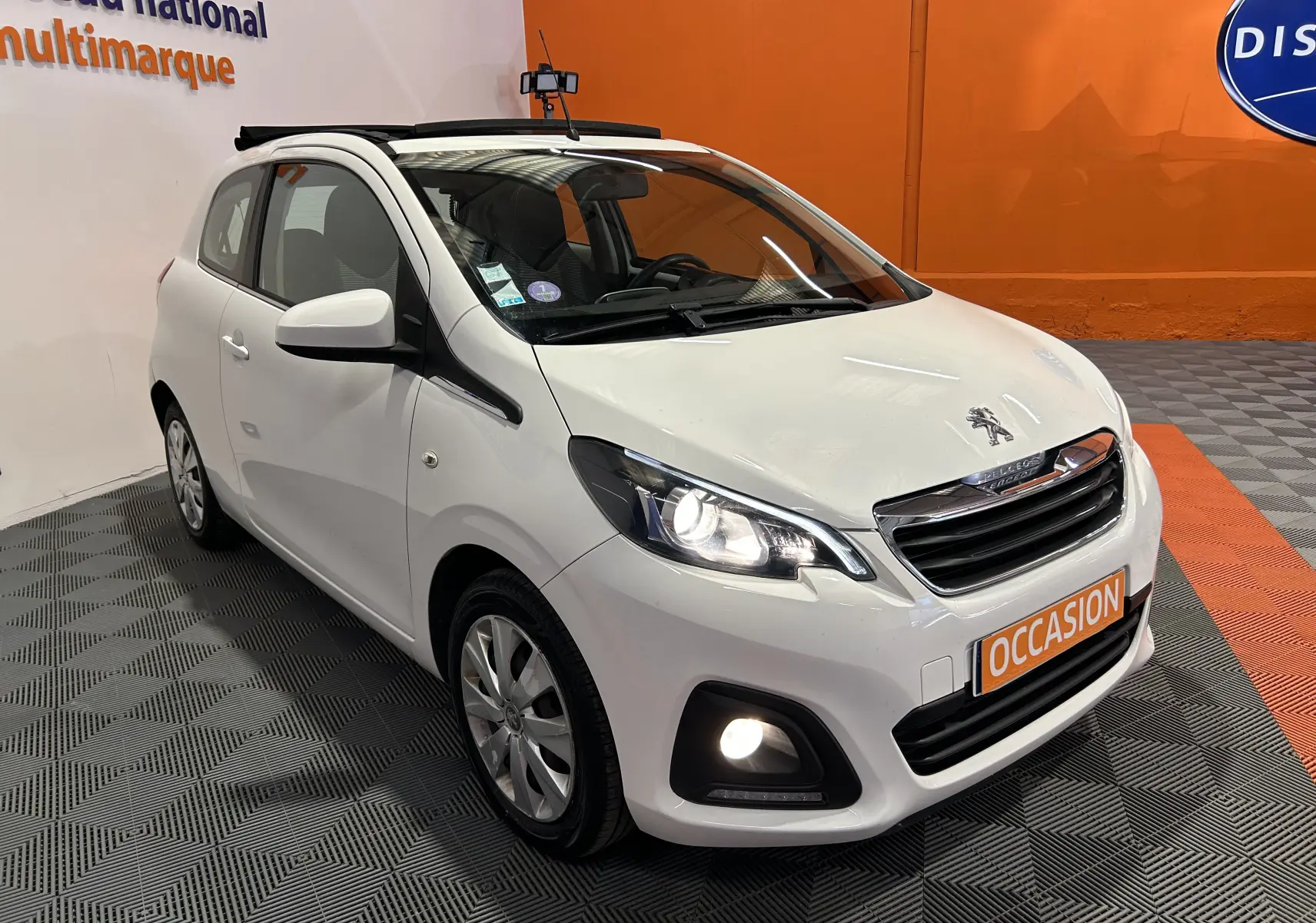 Vue 3/4 avant droite d'une Peugeot 108 blanche avec phares allumés et toit ouvrant, en showroom avec sol gris et orange.