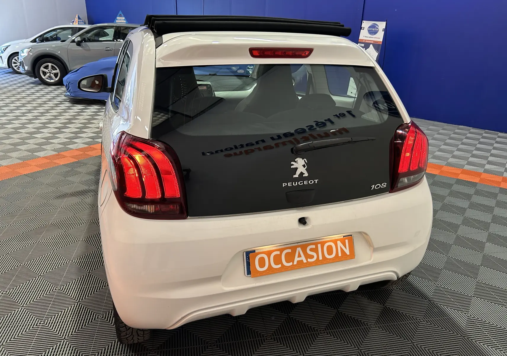 Vue arrière d'une Peugeot 108 blanche avec feux arrière allumés, affichant une plaque "OCCASION" en intérieur showroom.