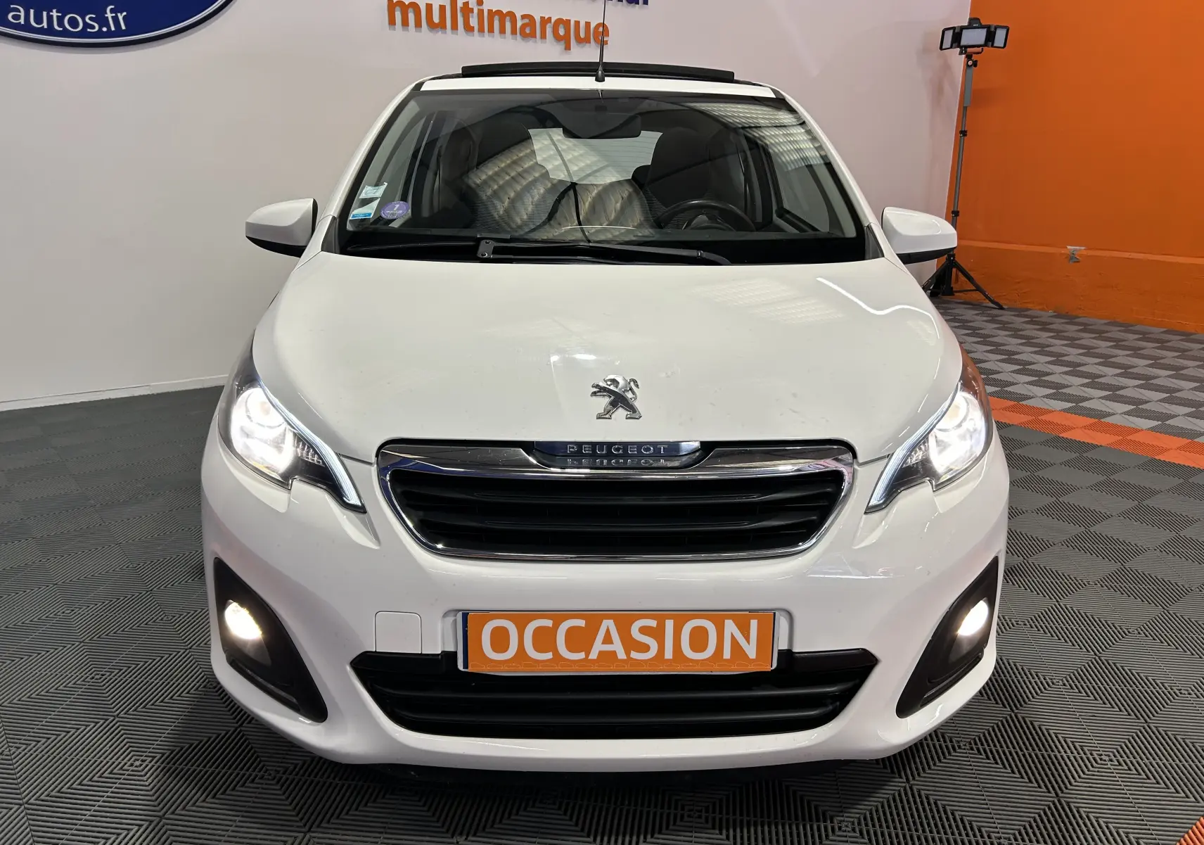 Vue avant d'une Peugeot 108 blanche avec phares et feux antibrouillard allumés dans un showroom.