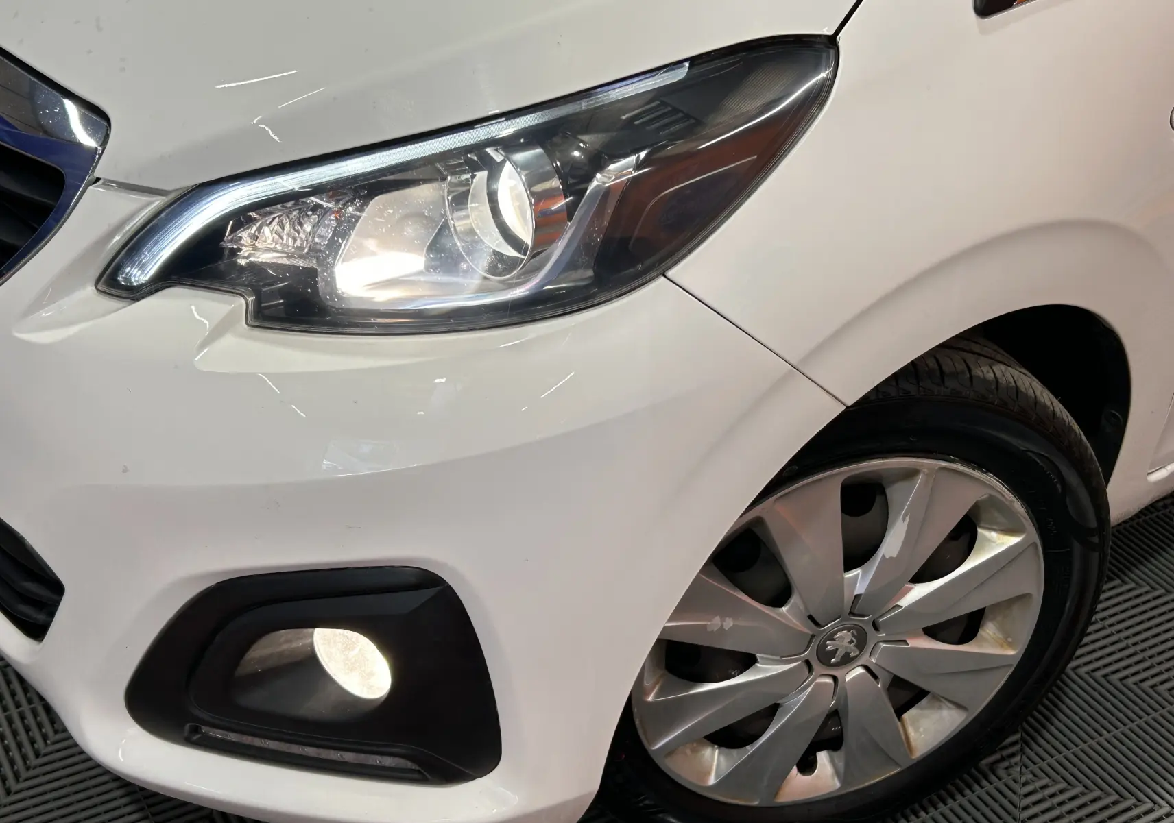 Gros plan sur l'avant droit blanc d'une Peugeot 108 2014, mettant en valeur le phare allumé et la roue avec enjoliveur.