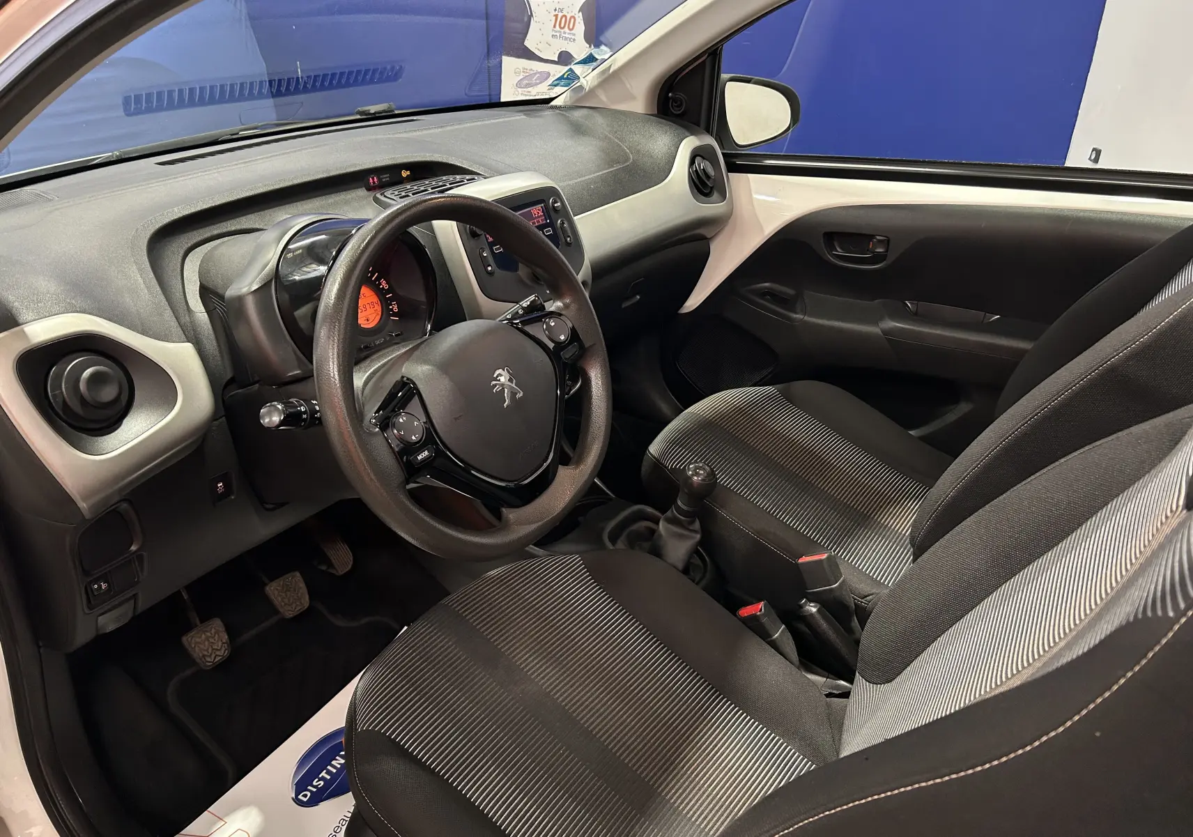 Vue intérieure côté conducteur de la Peugeot 108 blanche, montrant le volant, la planche de bord et les sièges tissu rayés noirs et gris.
