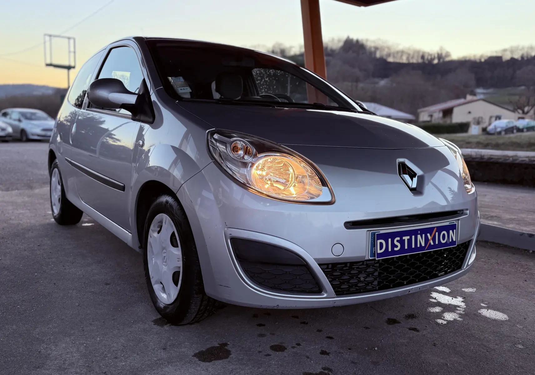 Renault Twingo II gris vue 3/4 avant droit, phare avant allumé sous un abri en fin de journée.