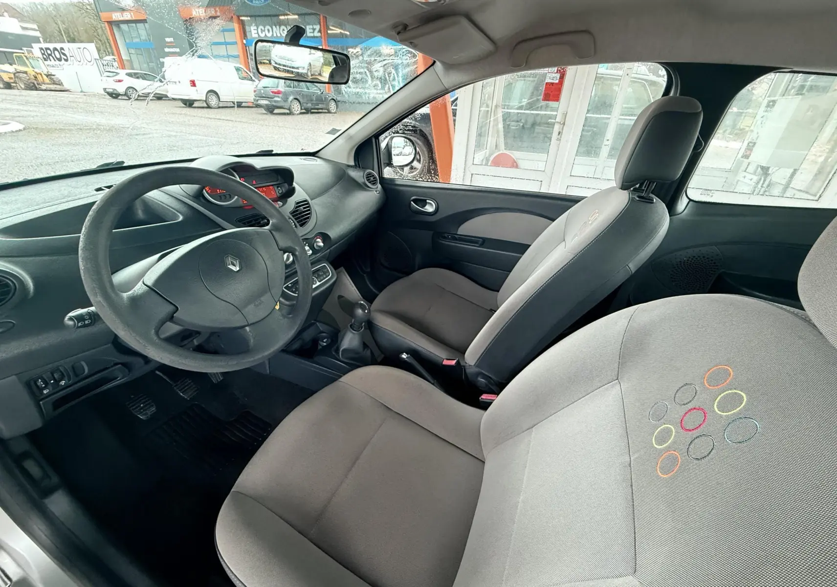 Vue intérieure côté conducteur de la Renault Twingo II 2009, sièges tissu gris avec motifs colorés et volant cuir noir.