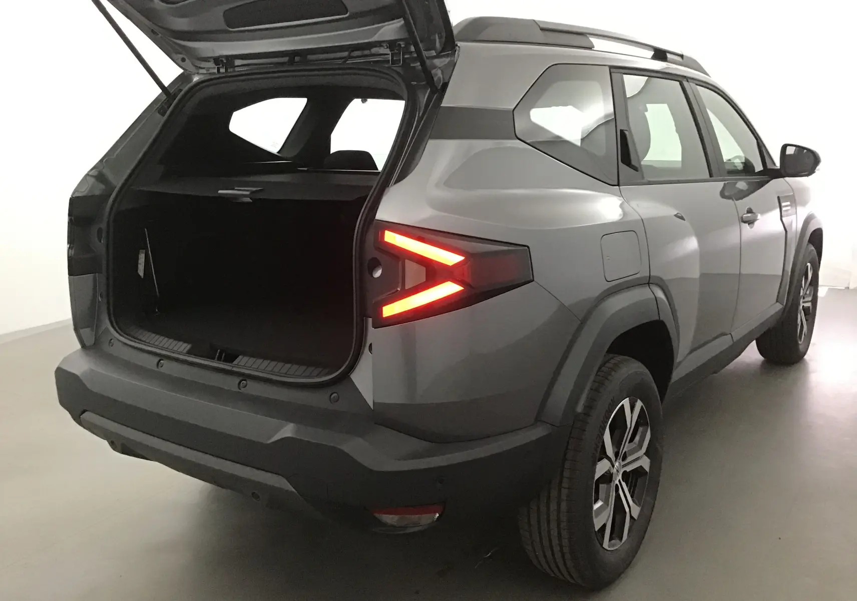 Vue 3/4 arrière droit du Dacia Bigster gris schiste avec coffre ouvert et feux arrière allumés.