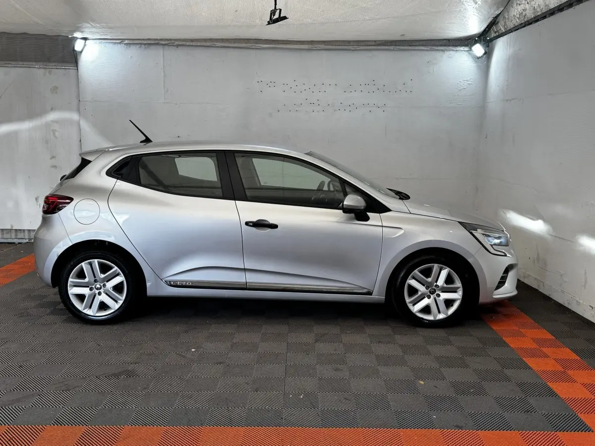 Profil droit d'une Renault Clio Business TCe 100 gris clair stationnée dans un garage avec sol noir et bandes orange.