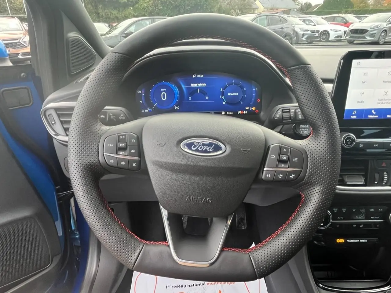 Vue intérieure centrée sur le volant perforé avec surpiqûres rouges du Ford Puma bleu 2022, tableau de bord numérique visible.