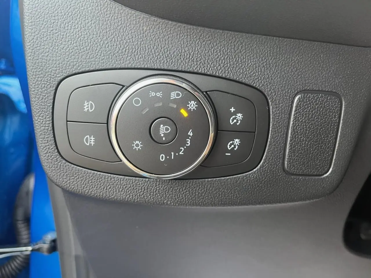 Gros plan sur le bouton de réglage des phares dans l'habitacle d'une Ford Puma bleu visible en arrière-plan.
