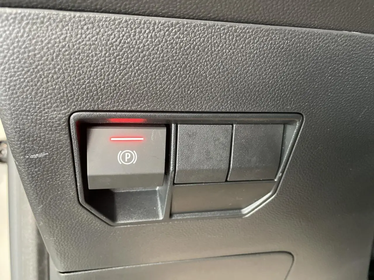 Gros plan sur le bouton de frein de stationnement électrique avec témoin rouge sur la console intérieure noire du Dacia Duster beige.