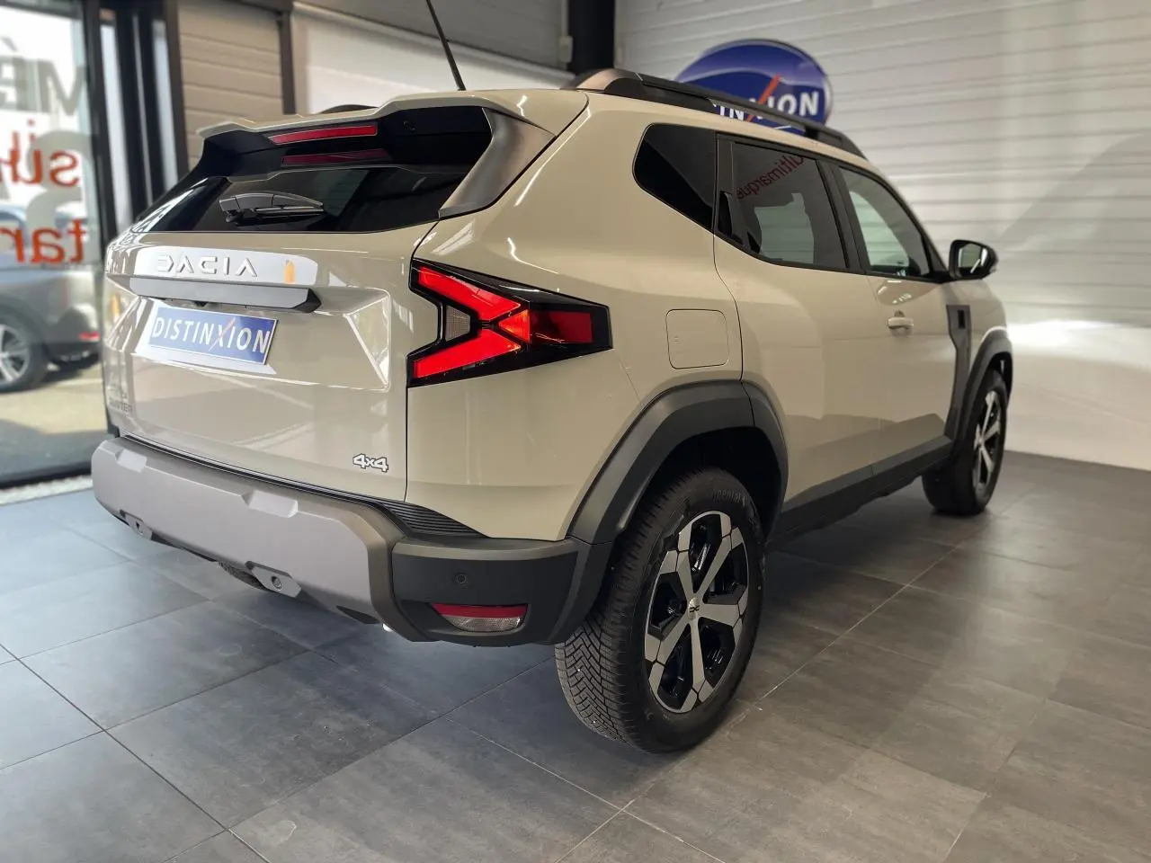 Vue 3/4 arrière droite du Dacia Duster beige 2025 avec jantes alliage noires et barres de toit noires.