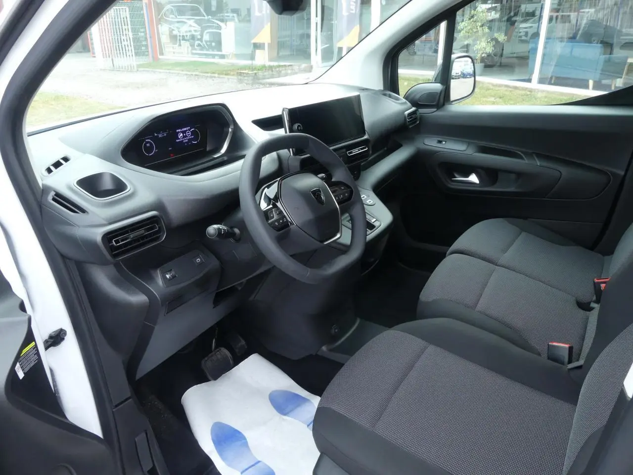 Vue intérieure côté conducteur du Peugeot Partner blanc 2025, avec tableau de bord numérique et sièges tissu gris.