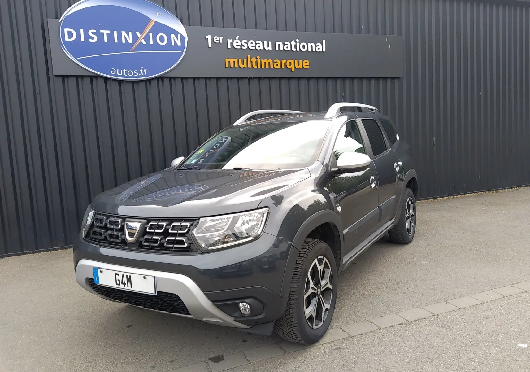 Dacia Duster gris 3/4 avant droit avec jantes alliage et barres de toit, devant un mur noir avec enseigne Distinxion.