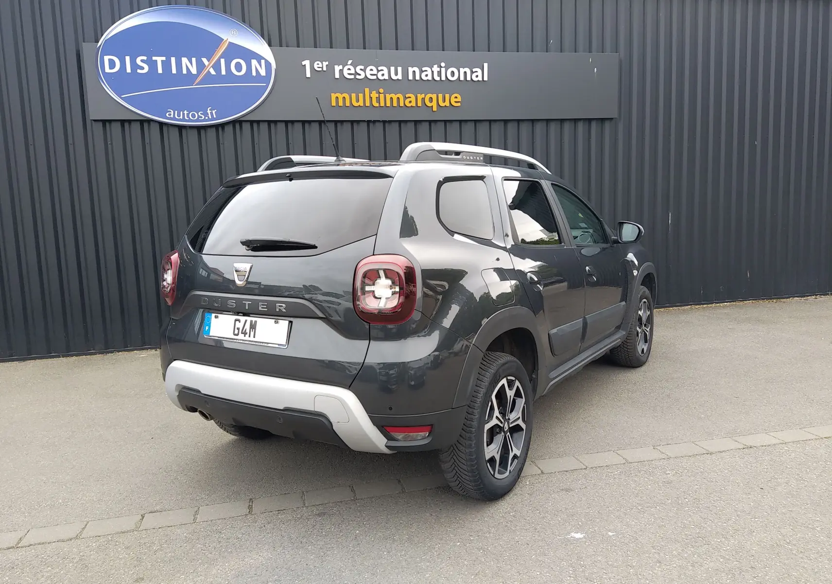 Vue 3/4 arrière droite d'un Dacia Duster gris 2020 avec jantes alliage et barres de toit, stationné devant un mur noir.