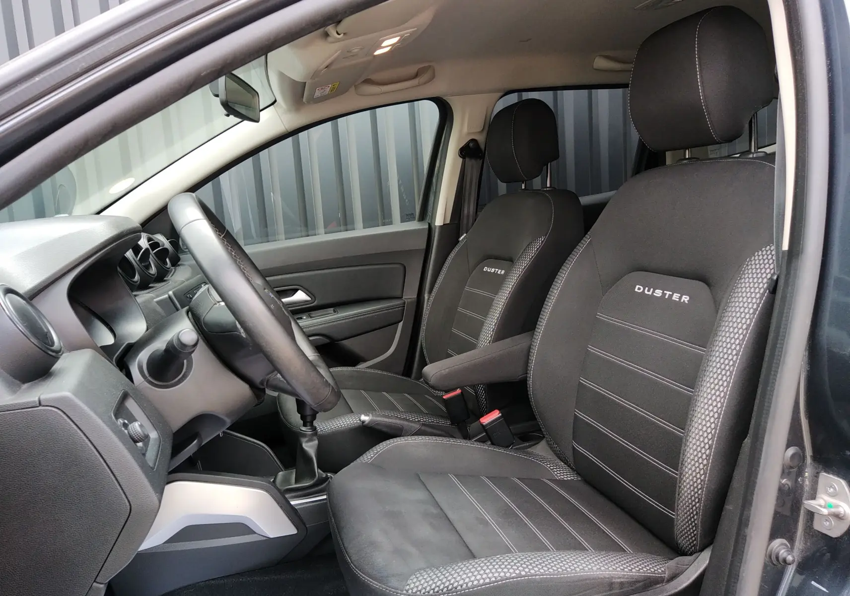 Vue intérieure côté conducteur du Dacia Duster gris 2020, sièges noirs avec logo Duster brodé et volant cuir.