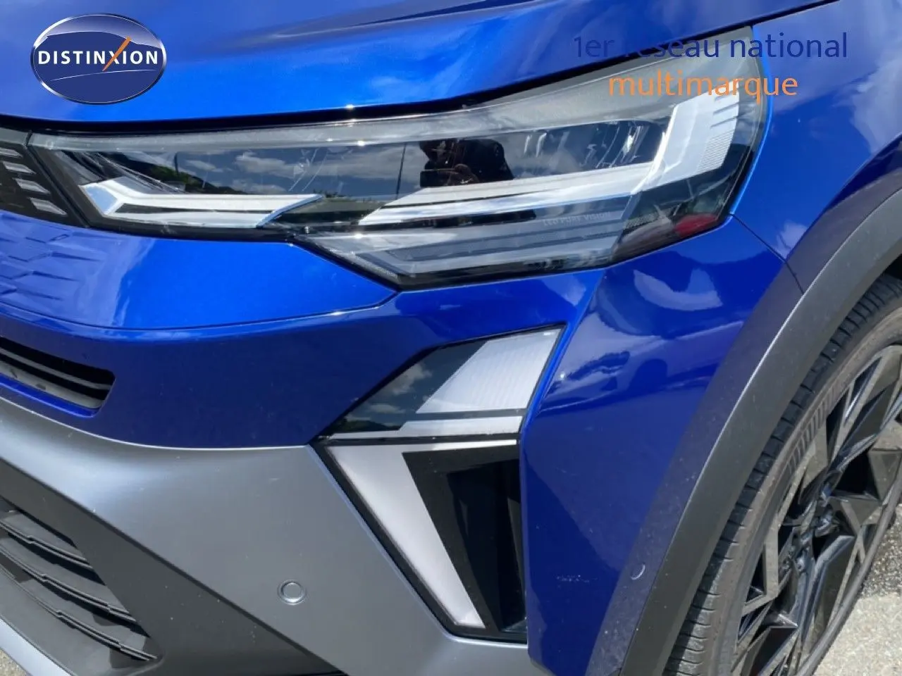 Gros plan sur l'avant droit bleu iron metal du Renault Symbioz E-Tech Full Hybrid, mettant en valeur le phare LED et la jante noire.