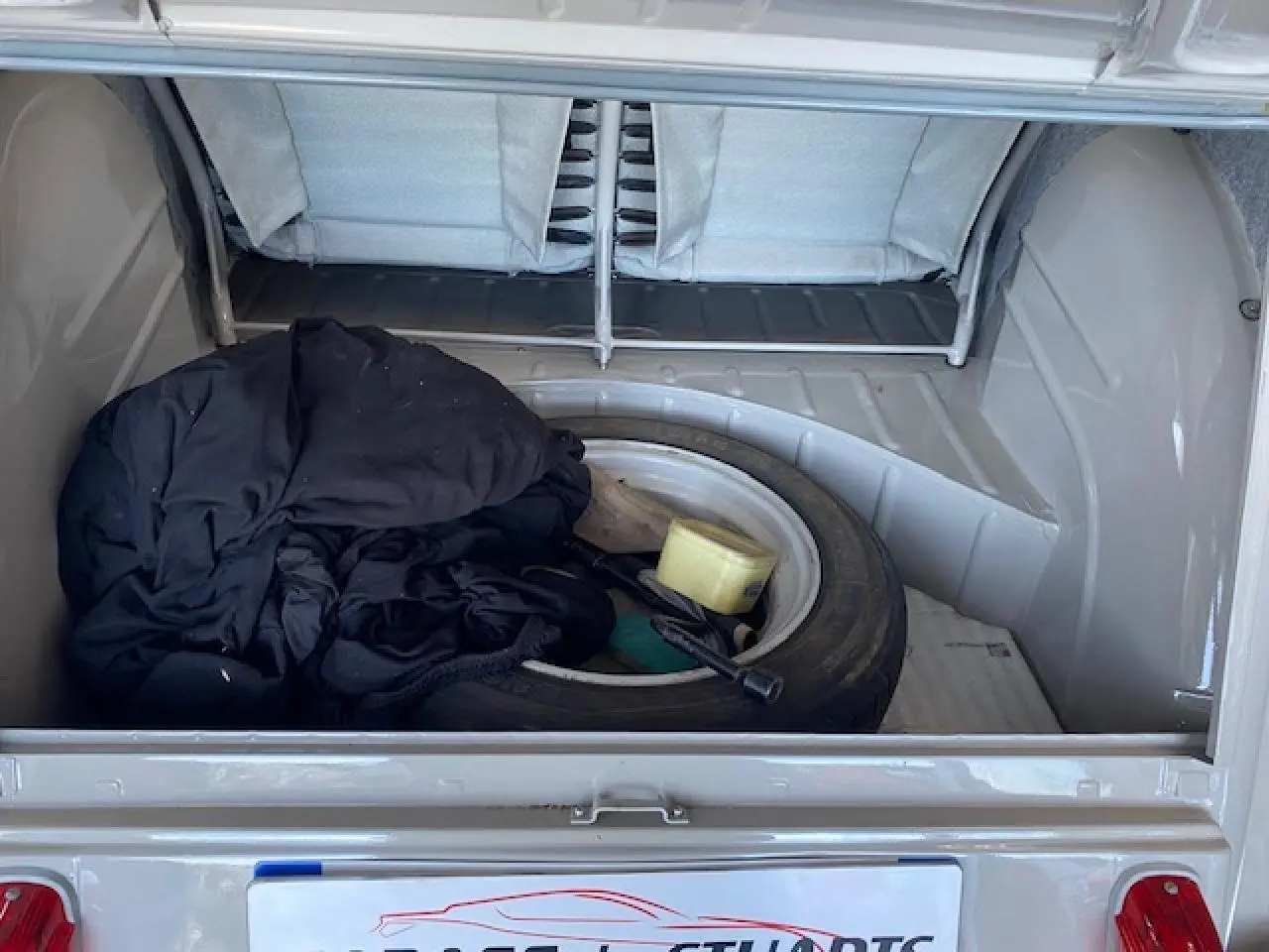 Coffre ouvert d'une Citroën 2 CV beige clair refaite à neuf, montrant une roue de secours et des objets à l'intérieur.