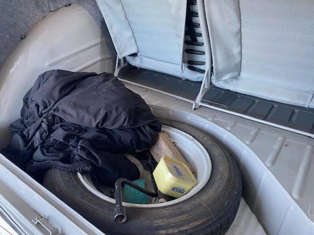 Coffre ouvert d'une Citroën 2 CV beige clair de 1968, montrant une roue de secours et des objets divers à l'intérieur.