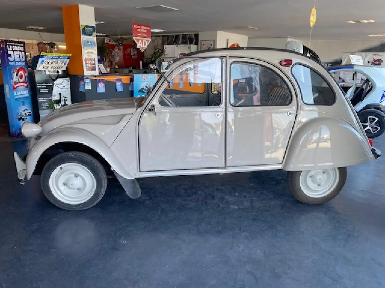 Profil côté gauche d'une Citroën 2 CV beige clair refaite à neuf, exposée en intérieur avec ses formes arrondies classiques.